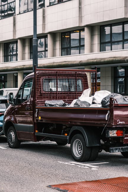 A small burgundy flatbed truck parked on an urban street in front of a modern office building with large glass windows and concrete cladding. The truck's open cargo area is filled with various waste m