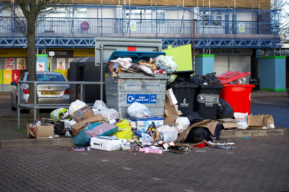 A collection of discarded rubbish including black plastic trash bags, which appear partially torn or opened, revealing assorted waste inside. There is a large, yellow plastic container lying on its si